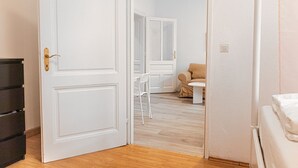 Una habitación con puerta blanca, suelo de madera, una silla blanca, una mesita pequeña de color blanco y una cama con ropa de cama blanca.