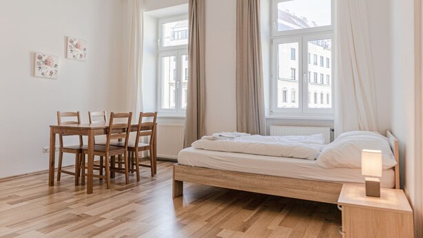 Un dormitorio con cama de madera, una mesita de noche con lámpara, una ventana con cortinas y una mesa de comedor con sillas.