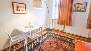 A room with a white table, two white chairs, a floor lamp, a patterned rug, a window with curtains, and two framed pictures on the wall.