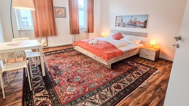 A bedroom with a bed, bedside tables, a desk, a chair, a large patterned rug, and a window with curtains.