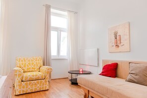 Una stanza con un divano, una poltrona a fantasia, un tavolino con una lampada, una finestra con tende e un quadro appeso al muro.