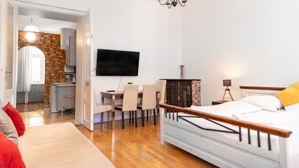A bedroom with a bed, a TV mounted on the wall, a dining area with chairs, and a kitchen visible through an open door. - Cheerful 2BR Apartment - Suitable for Longstays (Vienna)
