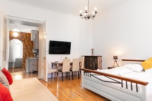 Una camera da letto con un letto, una TV fissata al muro, un'area pranzo con sedie e una cucina visibile attraverso una porta aperta.