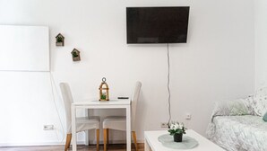 Une pièce avec un bureau blanc, deux chaises, une petite table avec une plante, un lit recouvert d’une couverture à motifs et un téléviseur fixé au mur.