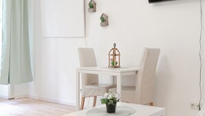 Une pièce au décor minimaliste, avec une table blanche, deux chaises, une petite plante en pot et un téléviseur fixé au mur.