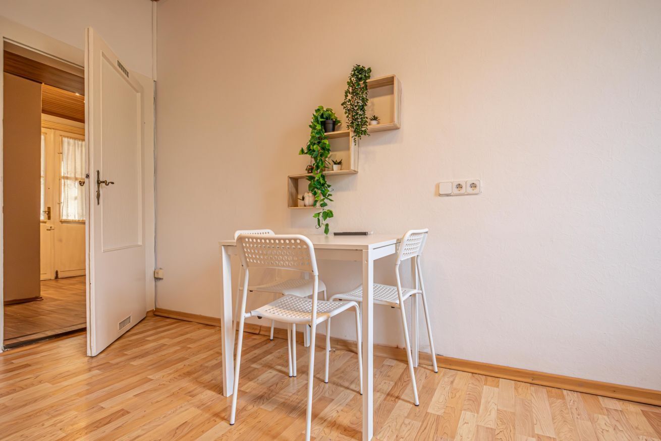 A room with a white table, two white chairs, a potted plant, and a door leading to another room.