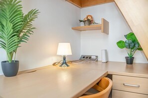 Une chambre moderne avec un bureau en bois, une lampe, une plante en pot, une étagère avec des fleurs et une station de recharge.