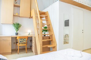 Une chambre avec un bureau en bois, une chaise, un lit, une échelle en bois, une plante en pot et un miroir.