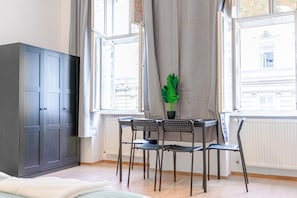 A room with a bed, a wardrobe, a table with chairs, a potted plant, and large windows.