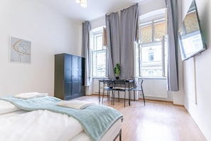 Une chambre avec un lit, un bureau et une chaise. Il y a une fenêtre avec des rideaux, un radiateur et une plante sur le bureau.