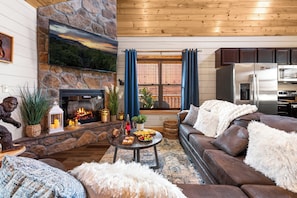 A cozy living room with a stone fireplace, a flat-screen TV, a modern kitchen, and a comfortable sofa.