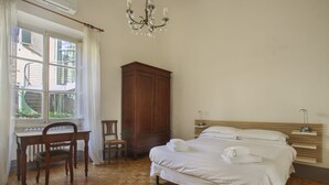 A bedroom with a wooden floor, a desk with a chair, a wardrobe, a bed with pillows, and a chandelier. - Lemon Tree One Bedroom with Balcony (Florence)