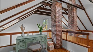 Une chambre à coucher de style loft avec des poutres en bois apparentes, une cheminée en briques, un bureau avec des tiroirs, une fenêtre et une vue sur l’extérieur.