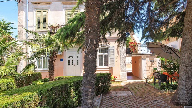 A building with a symmetrical facade, a central entrance with a door, and a small garden area with a palm tree and a paved walkway. - Holiday apartment with air conditioning (Fažana)