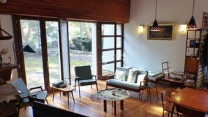 Ein geräumiges Wohnzimmer mit Holzboden, einer Couch, Sesseln, einem Couchtisch und einem Esstisch. Große Fenster sorgen für natürliches Licht und einen Ausblick ins Freie.