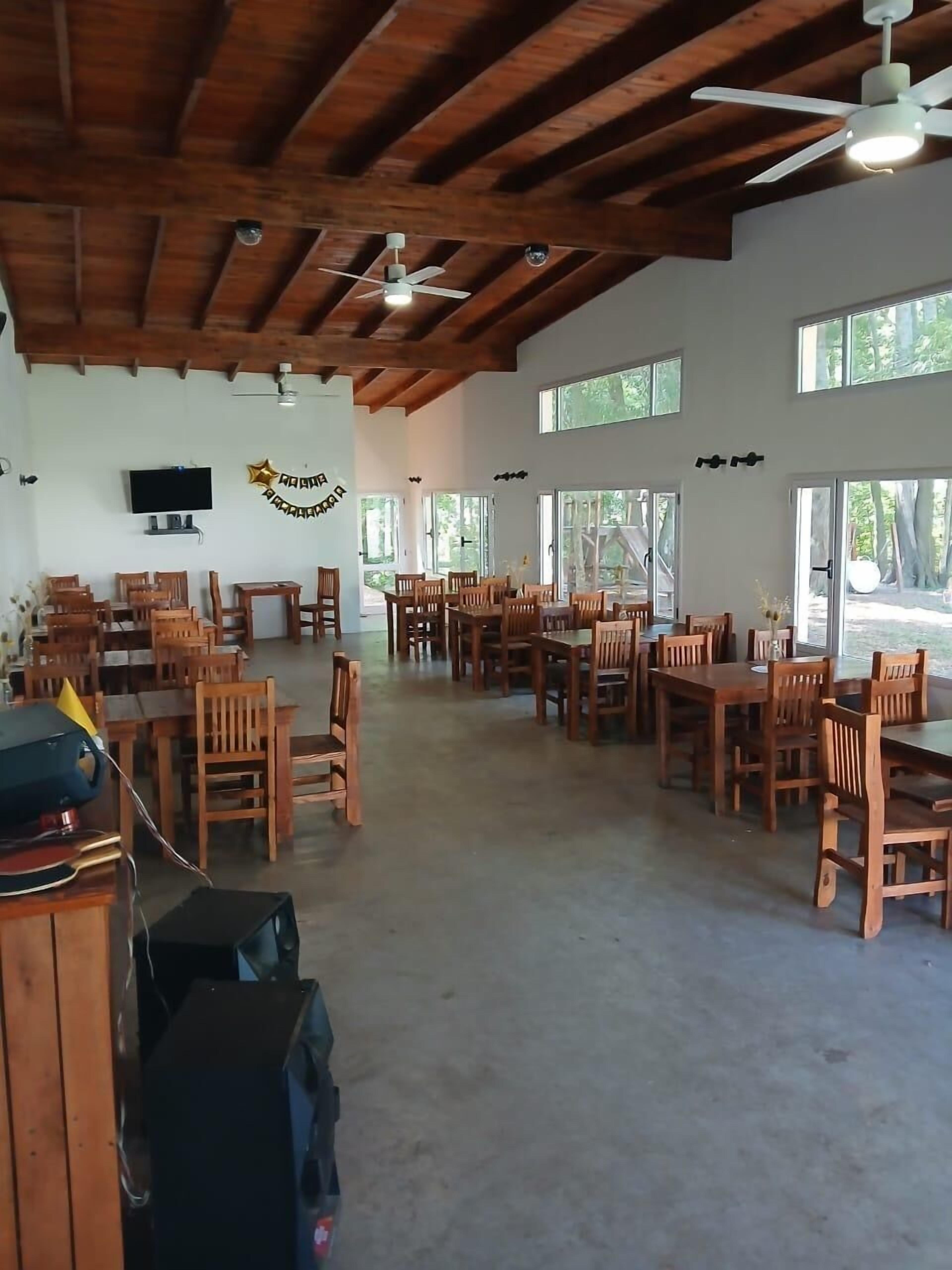 A spacious indoor area with wooden tables and chairs, ceiling fans, and large windows allowing natural light.