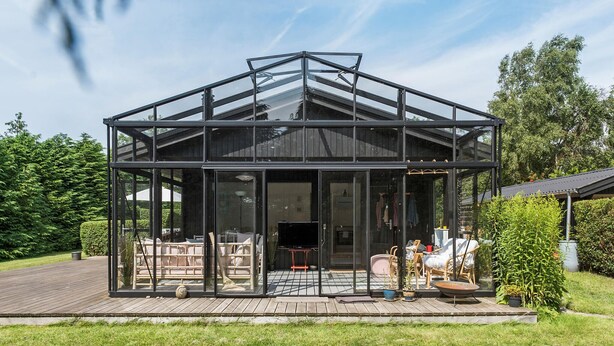 A modern glass house with a wooden deck, surrounded by greenery.