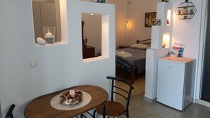 A compact hotel room with a dining area, a bed with a grey bedspread, a small refrigerator, and a wall-mounted shelf with decorative items. - Studio 'Gorgona' with sea view, Wi-Fi and air conditioning (Loutra)