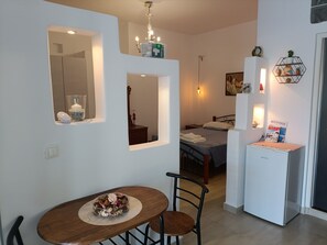 A compact hotel room with a dining area, a bed with a grey bedspread, a small refrigerator, and a wall-mounted shelf with decorative items.