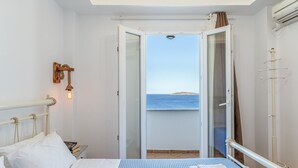 Ein Zimmer mit einem Bett, einem Nachttisch, einer Wandleuchte, einem Fenster mit Meerblick und einer offenen Tür, die auf einen Balkon führt.
