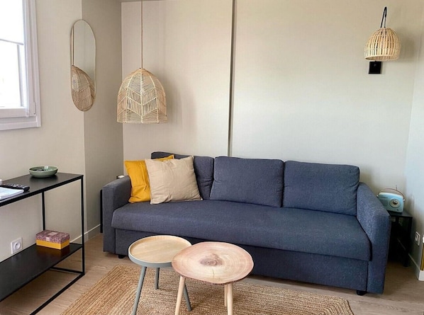 A modern living room with a dark blue sofa, two round wooden tables, a black side table, and a woven hanging light fixture.