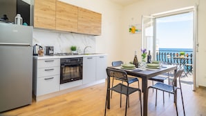 Una cocina moderna con horno integrado, un comedor con mesa y sillas, y vista al mar por una puerta abierta.
