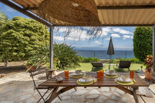 Un área de comedor al aire libre con una mesa preparada para cuatro, con vistas panorámicas del mar y las montañas.
