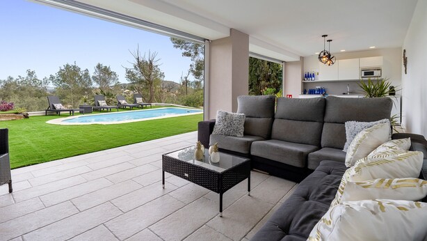 Un salon moderne doté d’une grande porte vitrée donnant sur une piscine, d’un canapé gris et d’une table basse en verre.