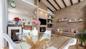 Une cuisine moderne avec des meubles blancs, un coin repas doté d’une table en verre et de chaises blanches, un mur en briques et une cheminée.