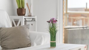 Une chambre avec un lit blanc, une table de chevet en bois ornée d’une plante en pot et un vase rempli de fleurs posé sur une table en bois.