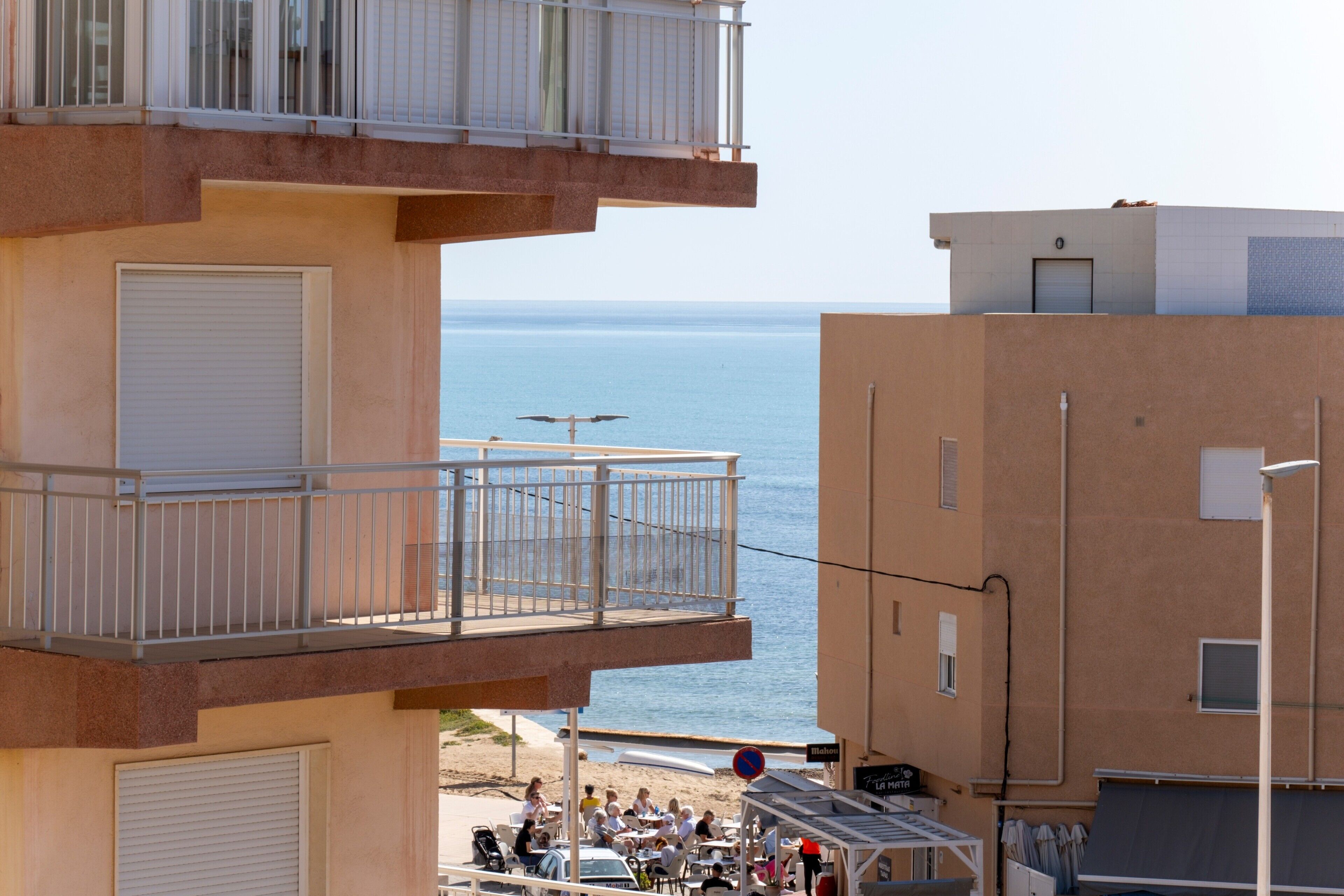 A building with a balcony overlooking the sea, a beach area with people, and a sign indicating a no parking zone.