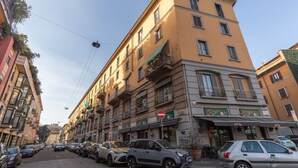 Eine Straße mit parkenden Autos und einem Gebäude mit mehreren Balkonen und Geschäften.