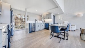 A modern kitchen with a central island, a dining area with blue chairs, and a large window overlooking a garden.