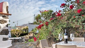 A table set with two wine glasses and a bottle, surrounded by wicker chairs, with a stone wall and red flowers in the background.