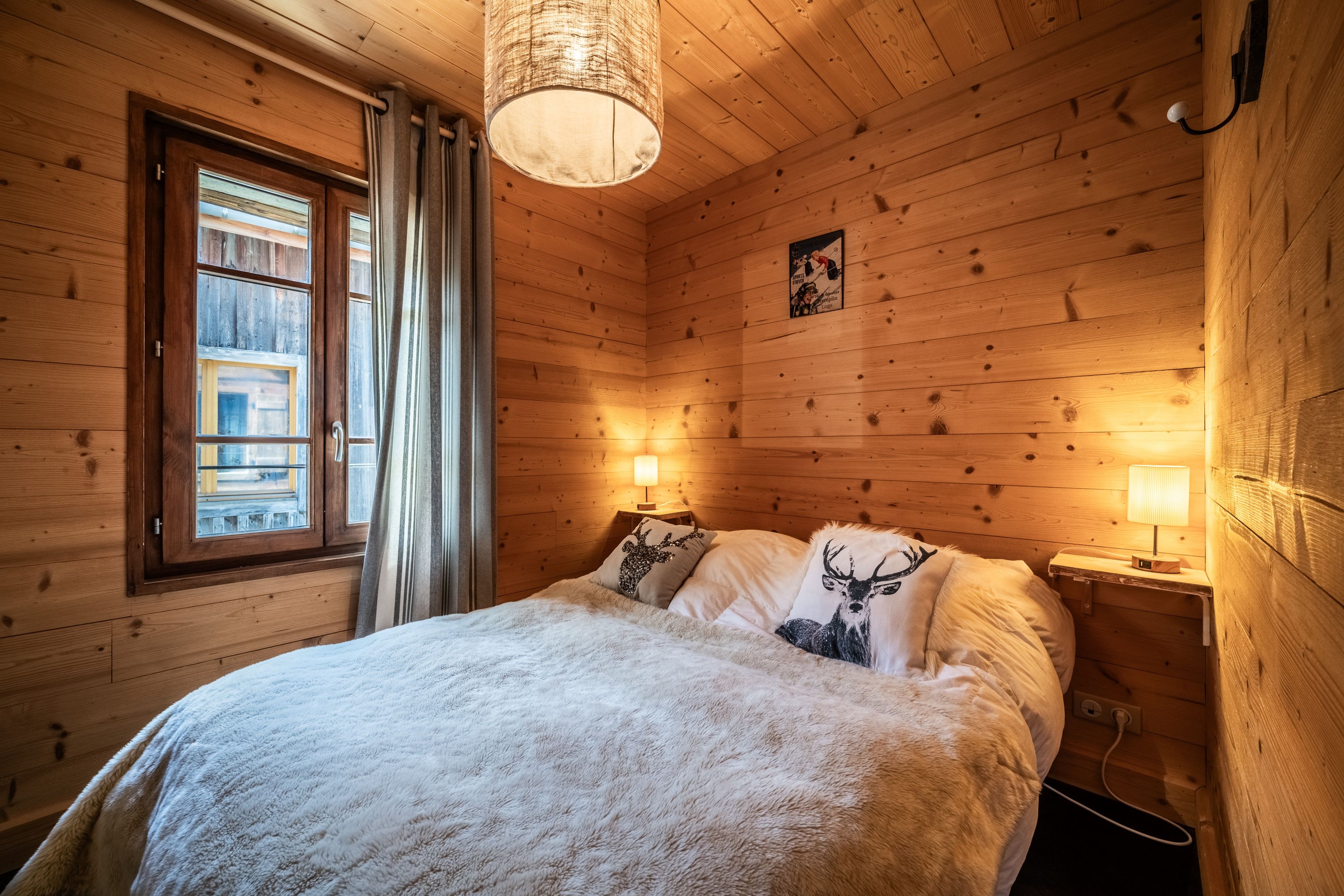 Ein Zimmer mit Holzboden, einem Bett, zwei Nachttischlampen, einem fenster mit Vorhängen und einer Wandleuchte.