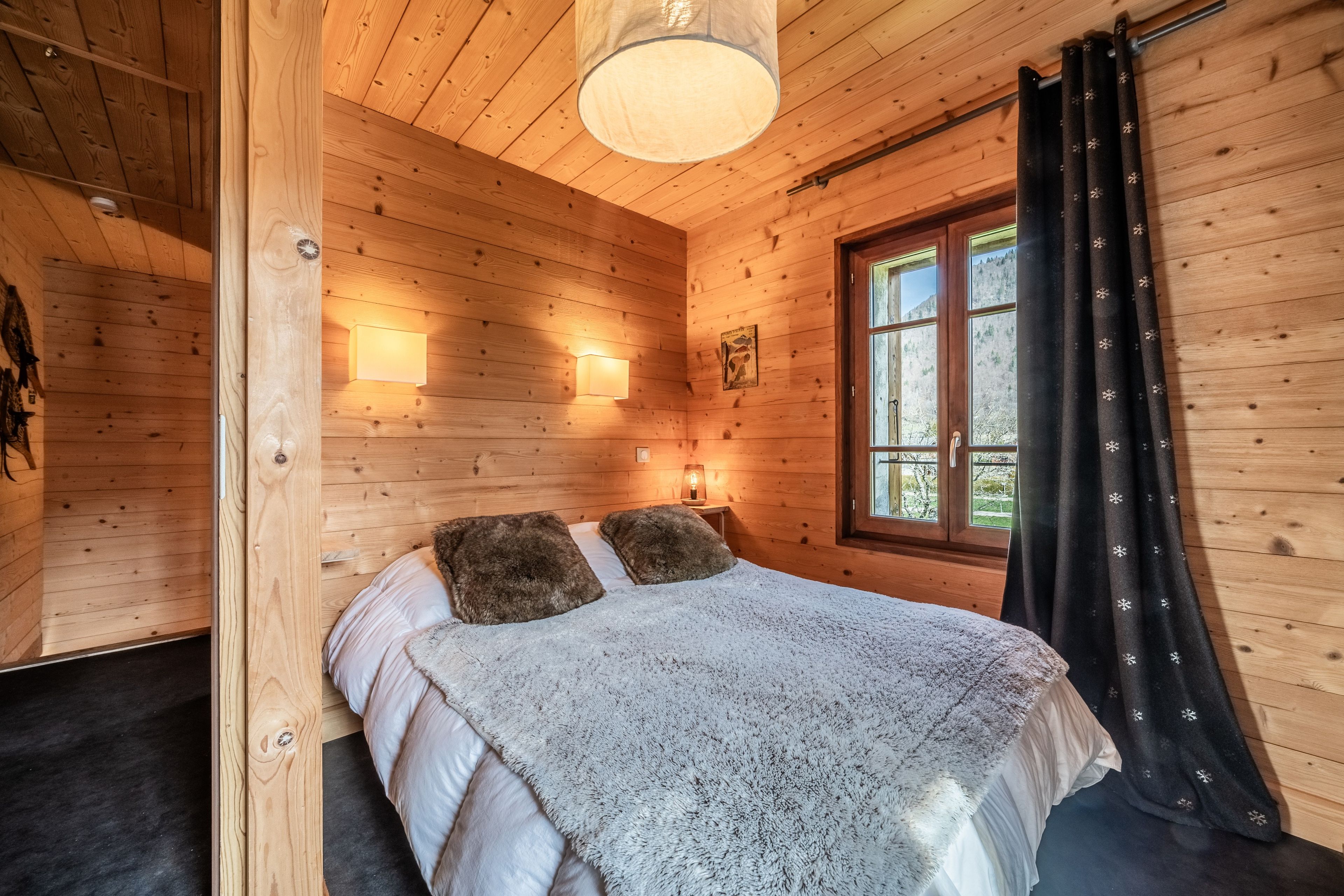 Ein Schlafzimmer einer Holzhütte mit einem Bett, zwei Wandleuchten, einem fenster mit Vorhängen und einer Hängelampe.