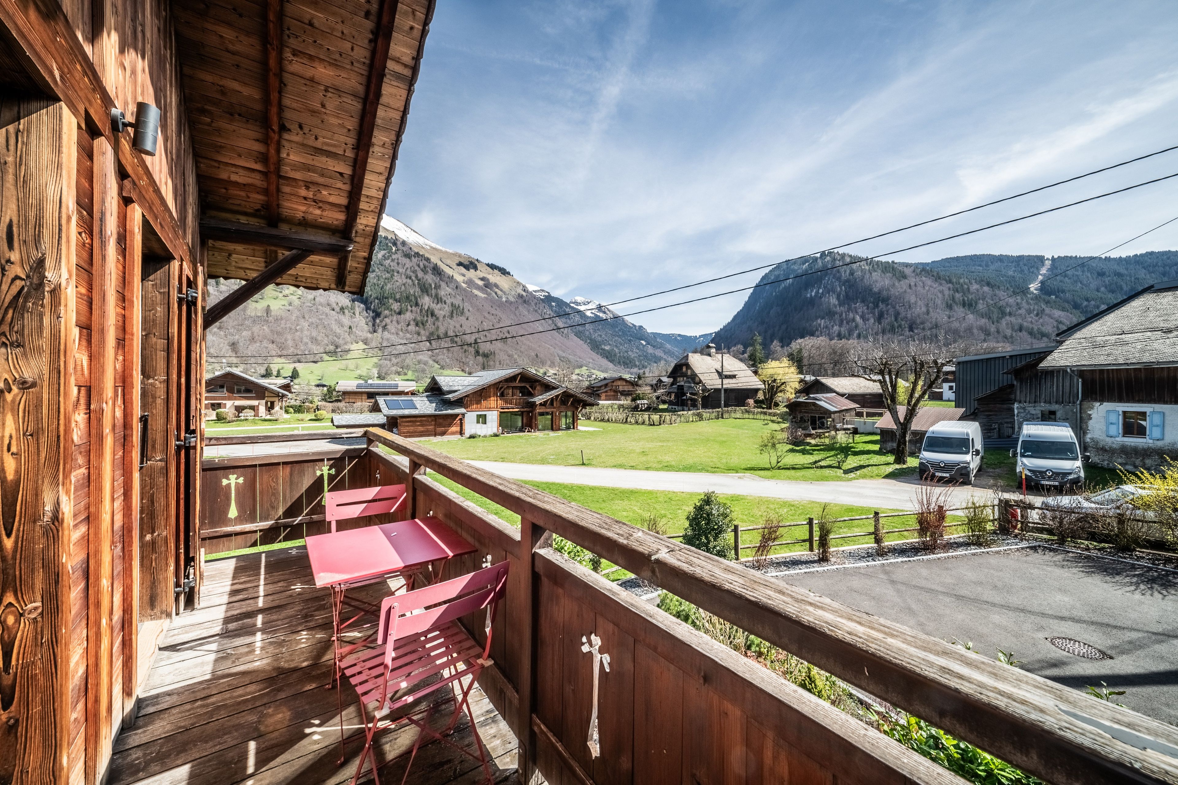 A wooden balcony with a table and chairs, overlooking a village with houses and mountains in the background.