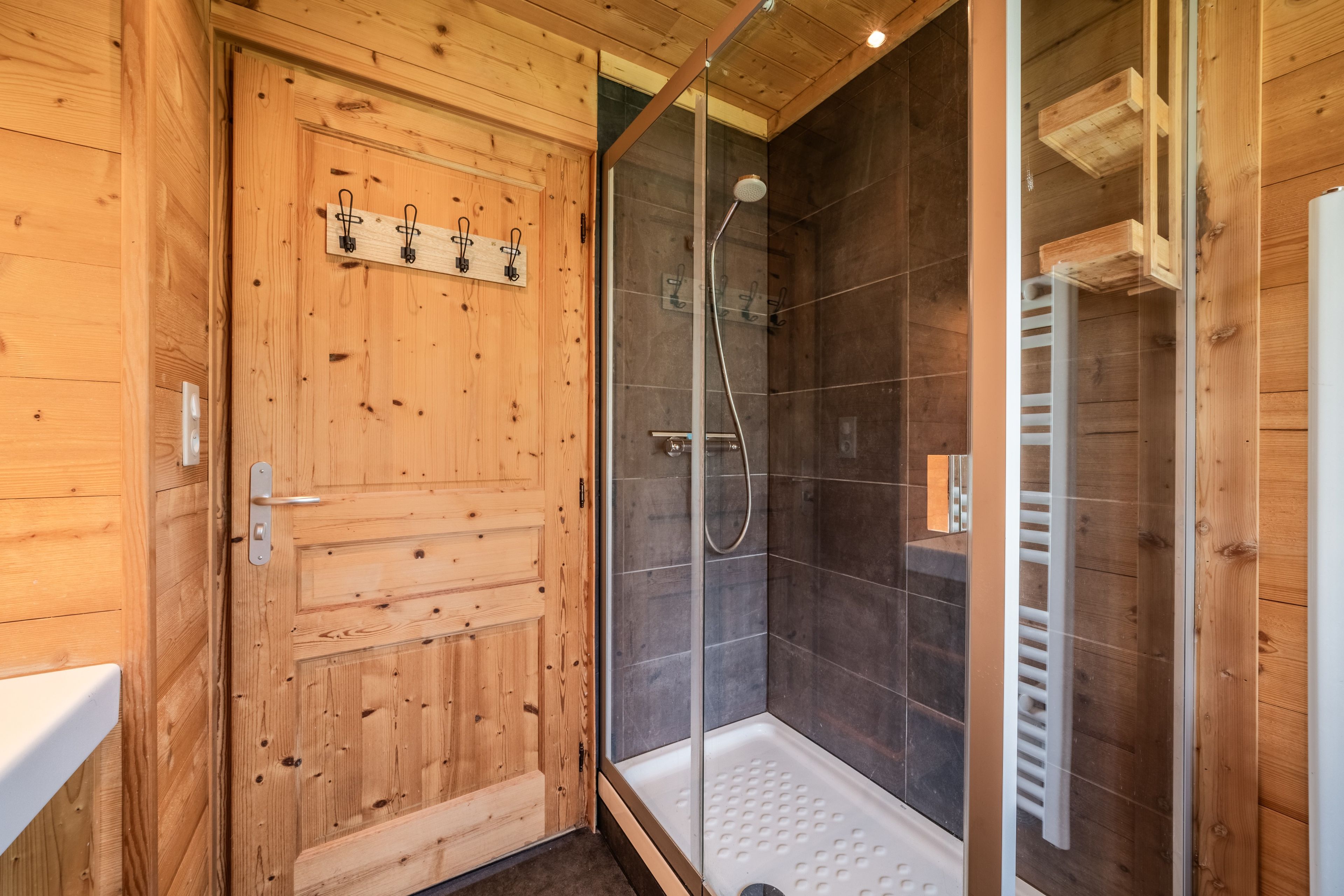 Interior de madera con cabina de ducha acristalada, puerta de madera con toallero y un lavamanos blanco.