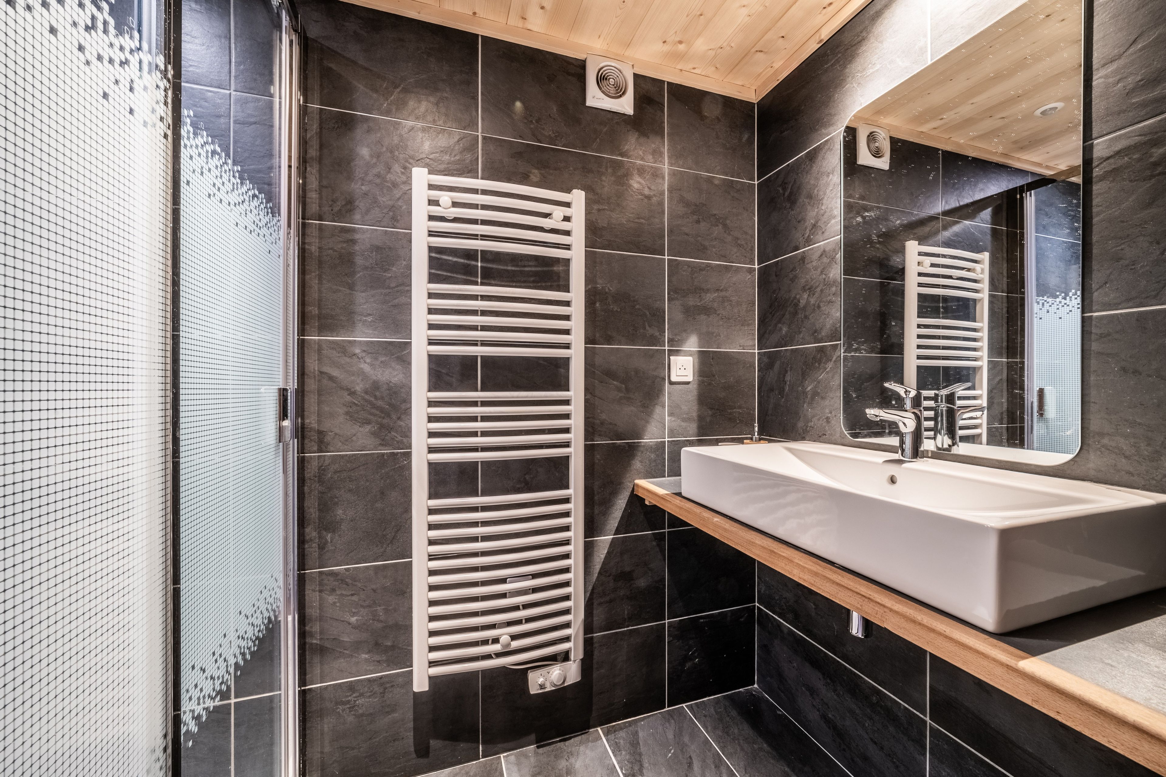 A modern bathroom with a white sink, dark tiled walls, and a wooden ceiling.