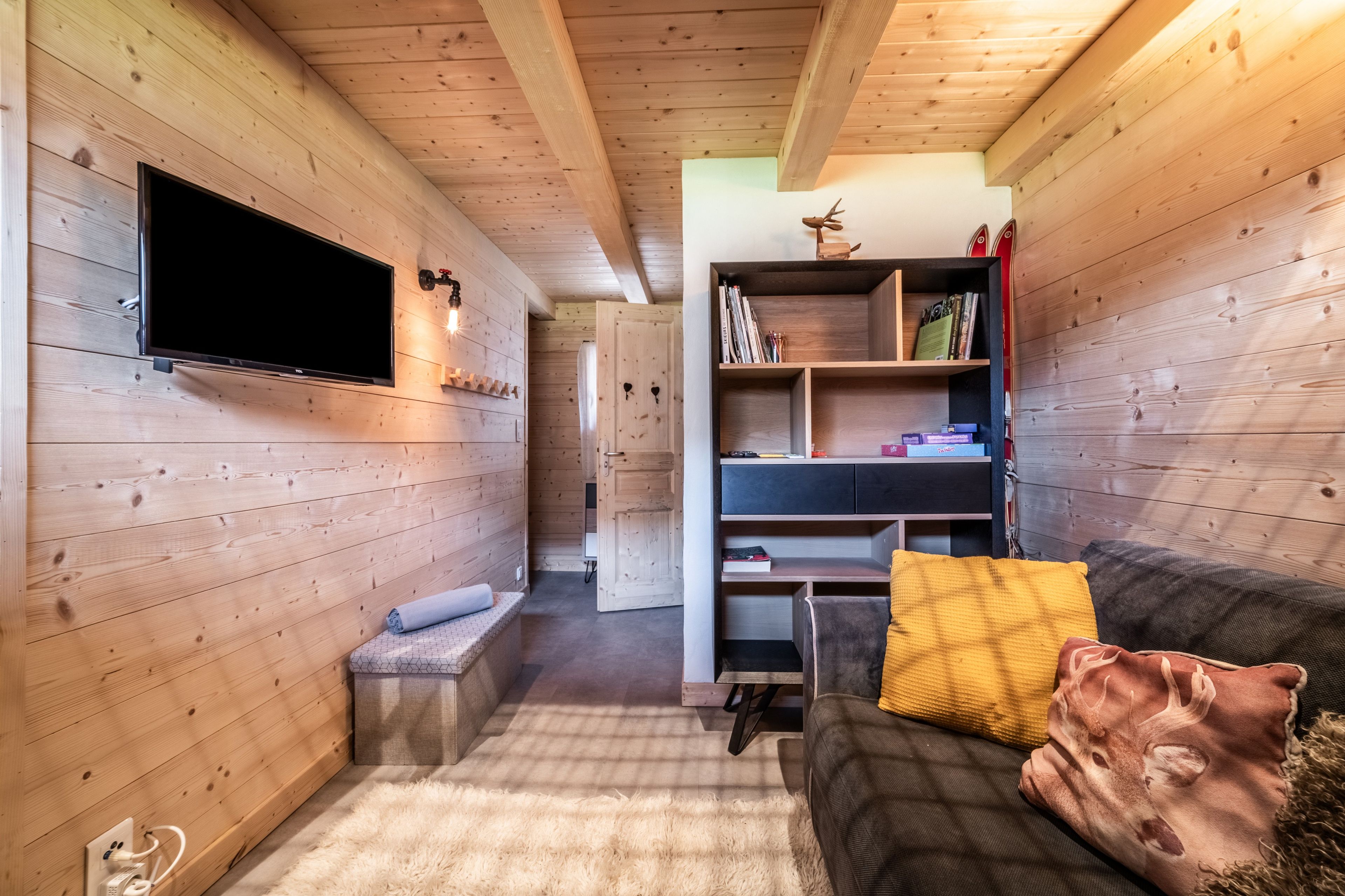 Une pièce confortable avec un téléviseur à écran plat fixé au mur, une bibliothèque en bois, un canapé avec des coussins et un petit banc.