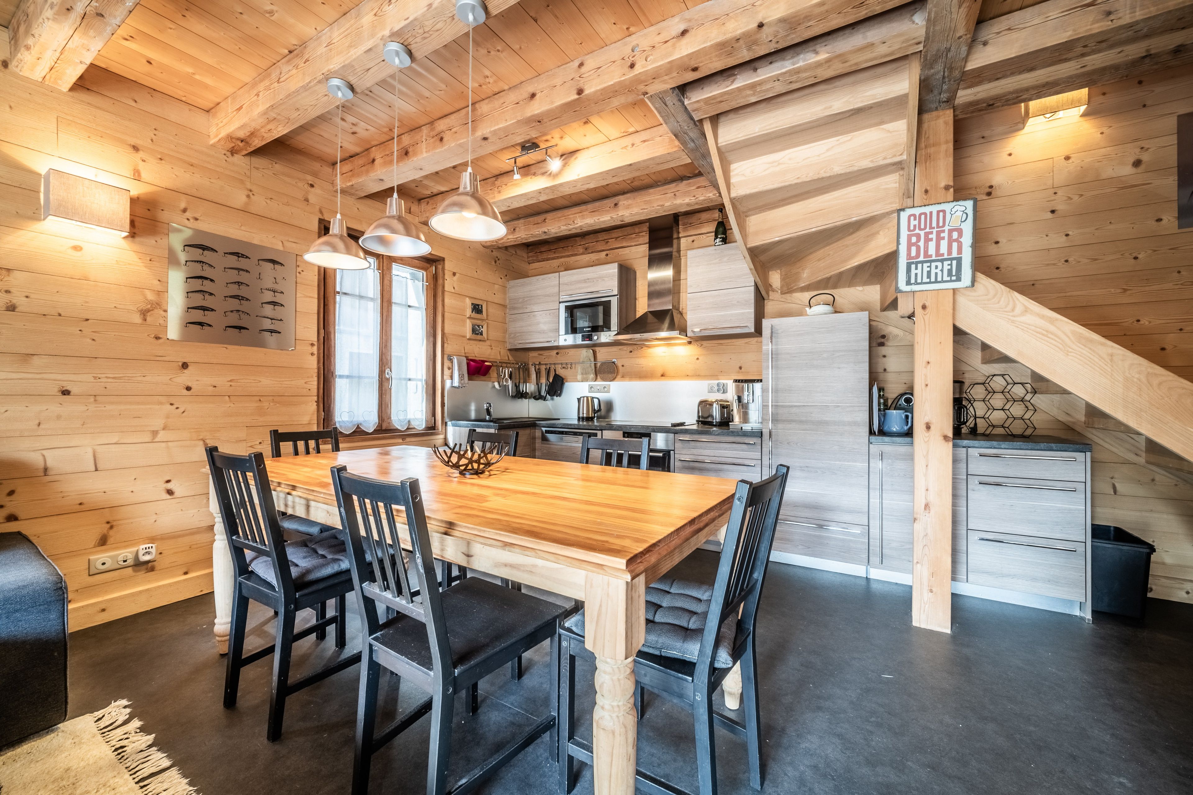 Een houten interieur met een eettafel, stoelen, een keukenhoek, en een bordje waarop 'koud biertje' staat.