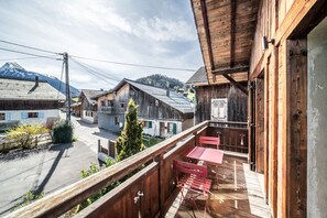 A wooden balcony with a red table and chairs, overlooking a street with traditional buildings and a mountain in the background.