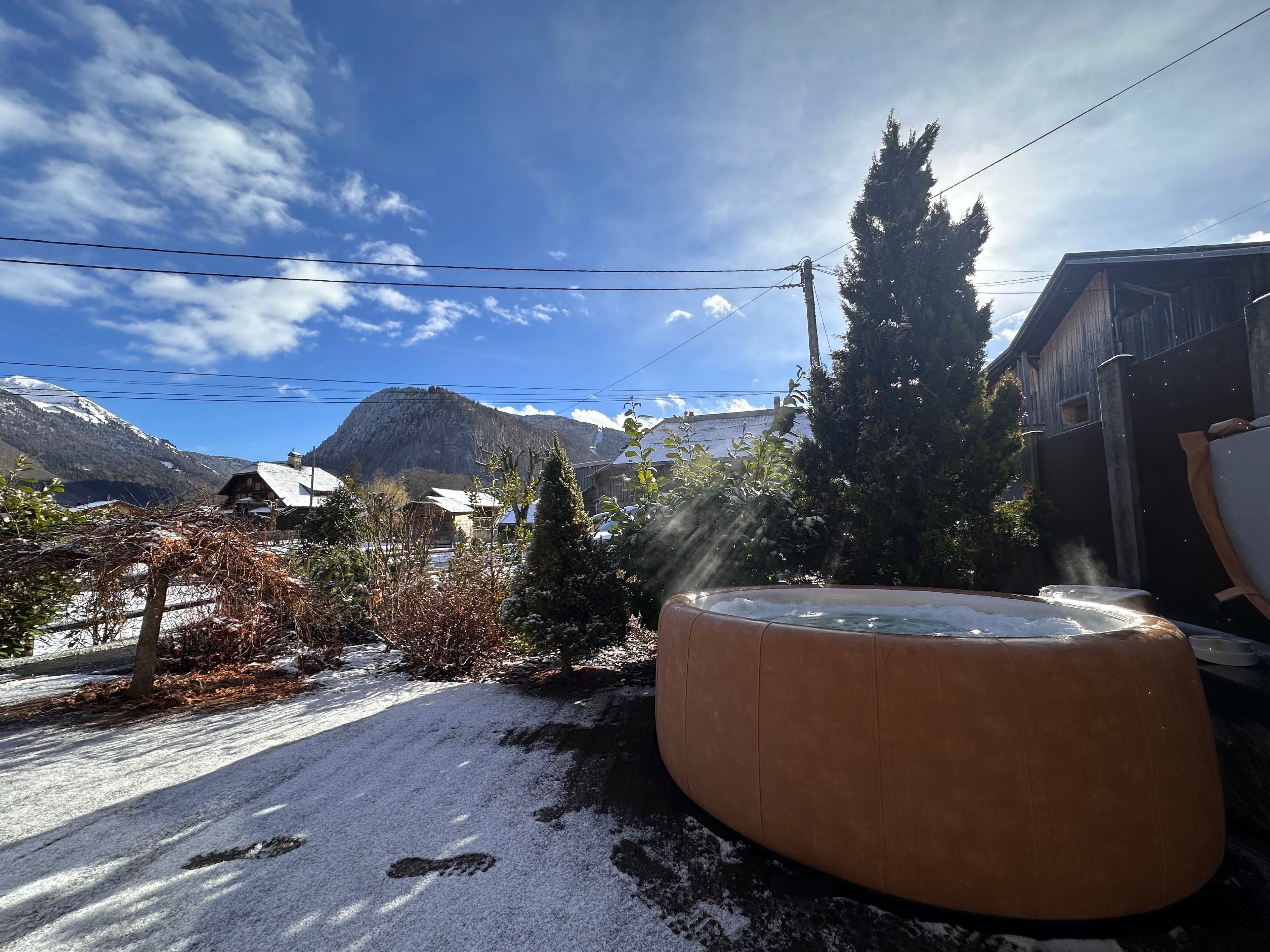 Un espace extérieur enneigé avec un jacuzzi, un bâtiment en bois et des montagnes en arrière-plan.
