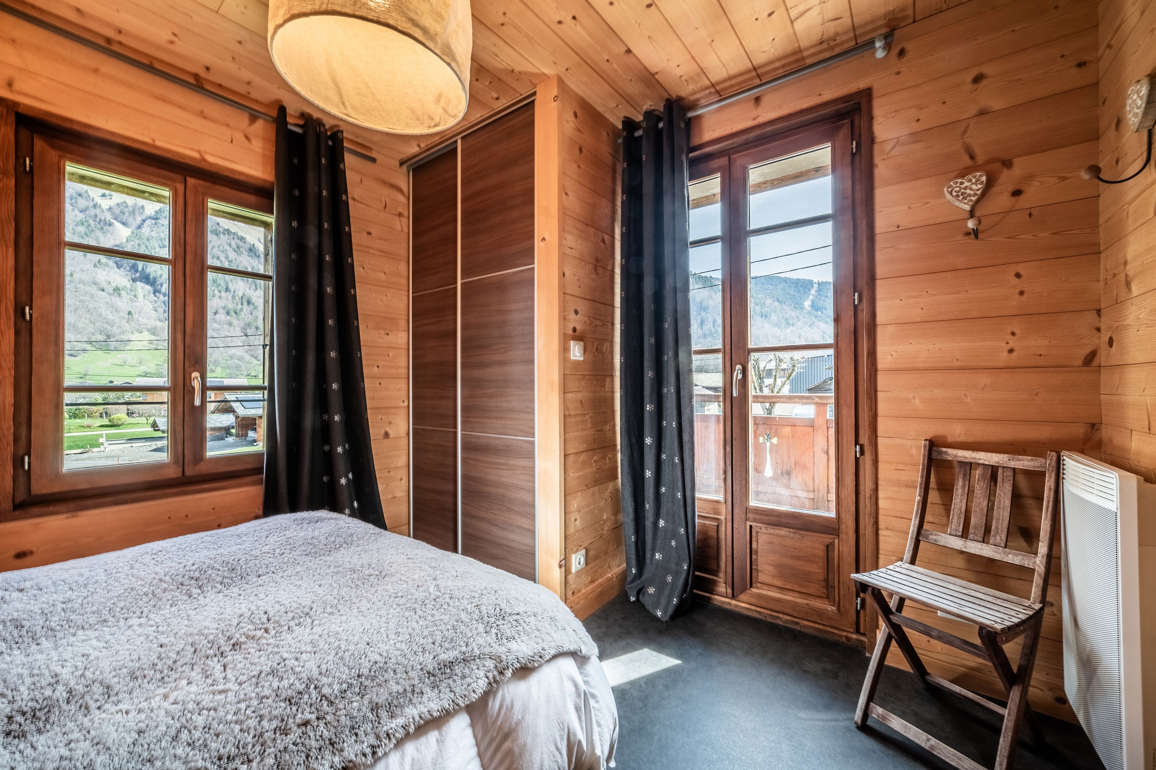 Ein Zimmer mit Holzboden, einem Bett, einem Sessel und Blick auf eine Berglandschaft durch die Fenster.