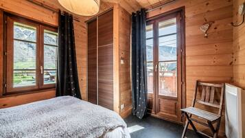Ein Zimmer mit Holzboden, einem Bett, einem Sessel und Blick auf eine Berglandschaft durch die Fenster.