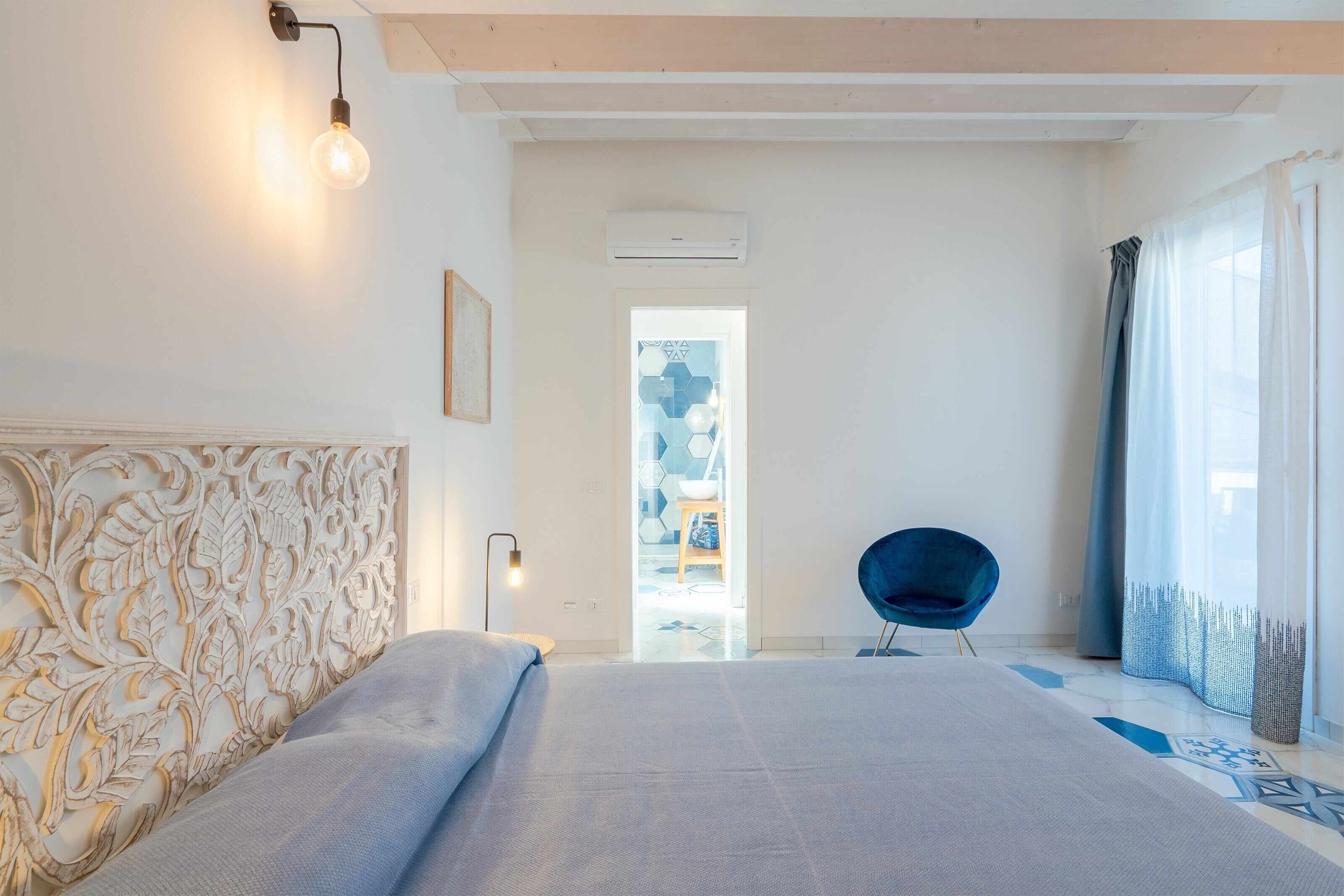 A modern bedroom with a large bed, a decorative headboard, a blue chair, and a view through a doorway.