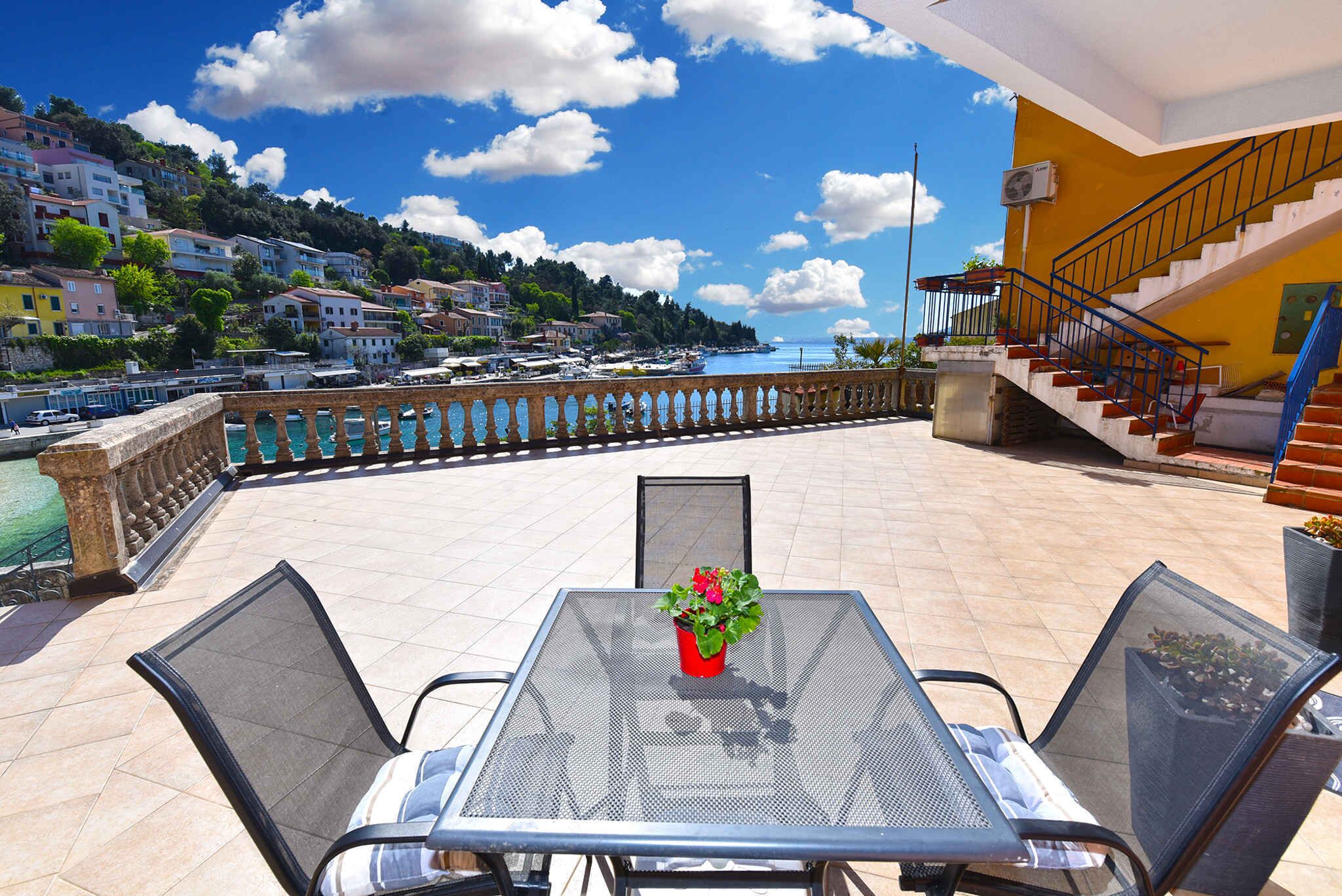 A terrace with a table and chairs, overlooking a coastal town with buildings and boats.