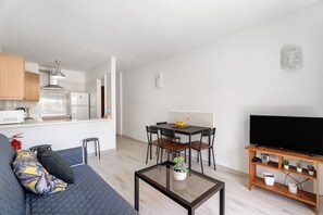 Un espace de vie moderne comprenant un canapé, une table à manger avec des chaises, un téléviseur posé sur un meuble en bois, et un coin cuisine à l’arrière-plan.