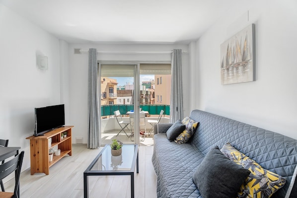 A modern living room with a sofa, a glass coffee table, a TV on a wooden stand, and a balcony with a view of buildings.