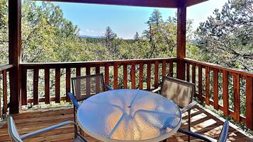 A covered outdoor deck with a round glass table and four chairs, surrounded by a wooden railing, overlooking a forested landscape.
